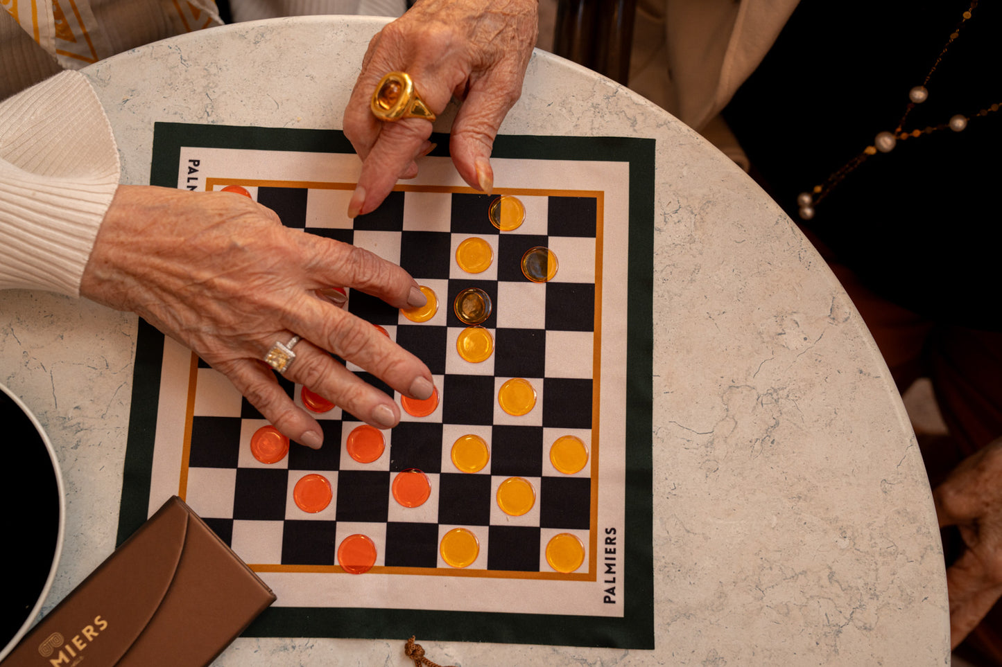 Older hands playing a game of checkers on a board cloth with checker pieces using colors of orange, yellow, green, black and ivory.